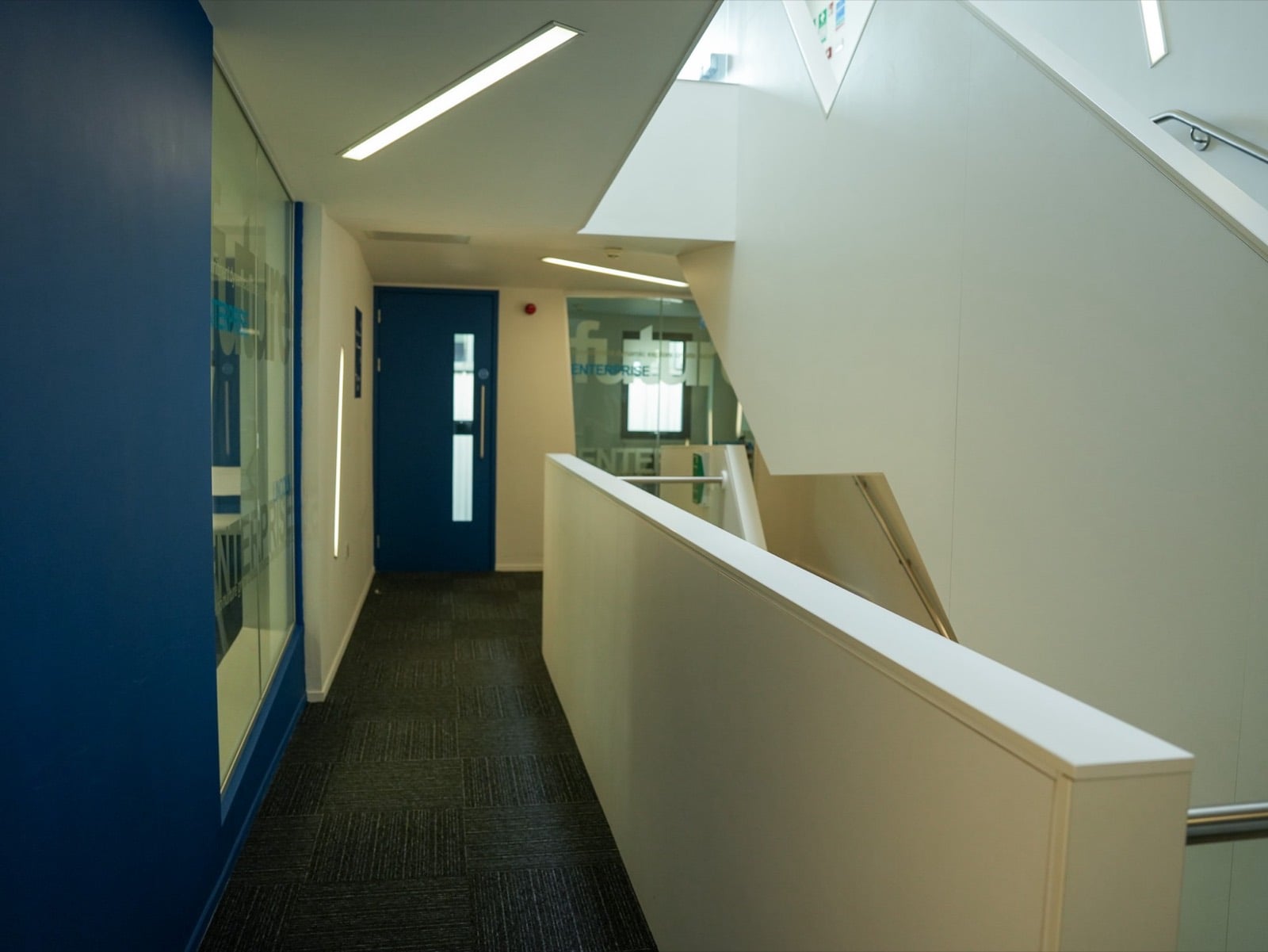 A corridor past a blue wall and glazed offices, with angled ceiling lights and the stairwell alongside