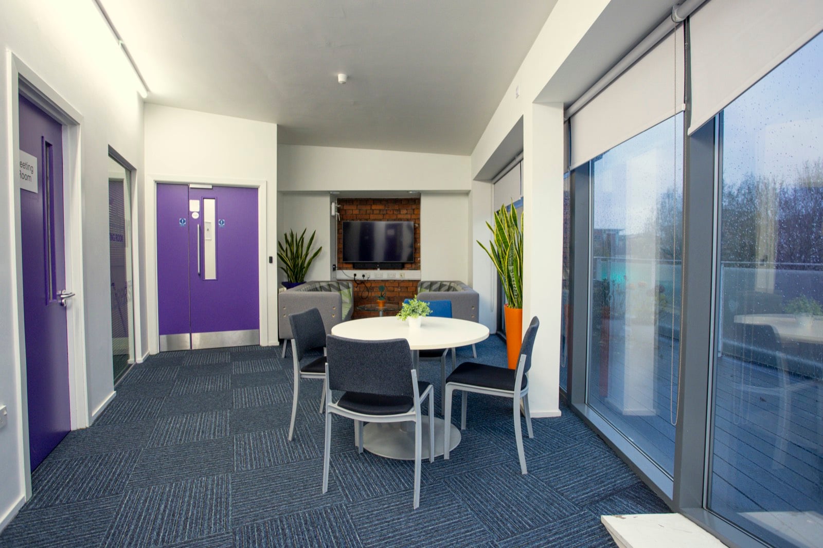 A shared breakout area with a round table, sofas and a screen, beside meeting room doors and a wall of glass onto the terrace