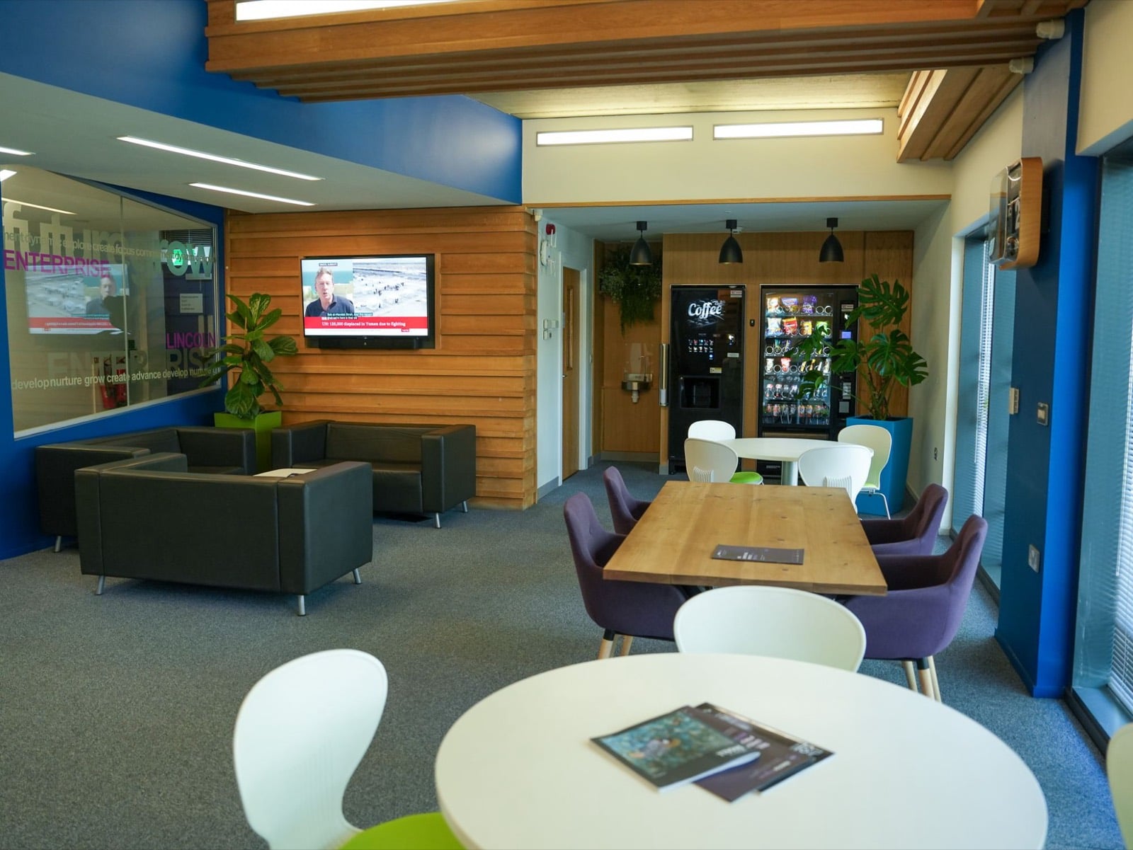 A shared lounge with leather sofas, a TV on a timber wall, coffee and snack machines, and tables by the windows