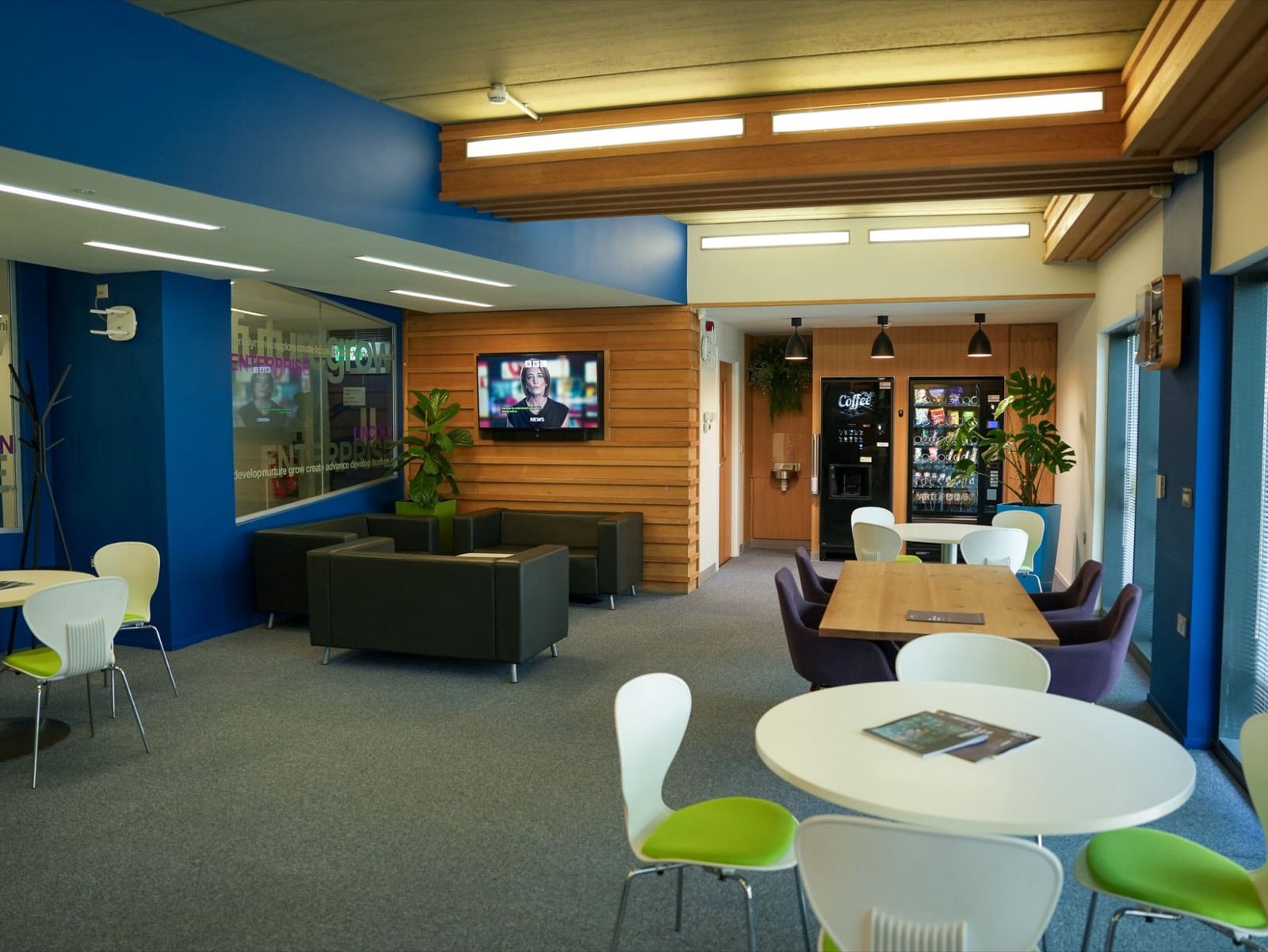 A shared lounge and cafe area with leather sofas, a wall-mounted TV, coffee and snack machines, and round tables with green chairs
