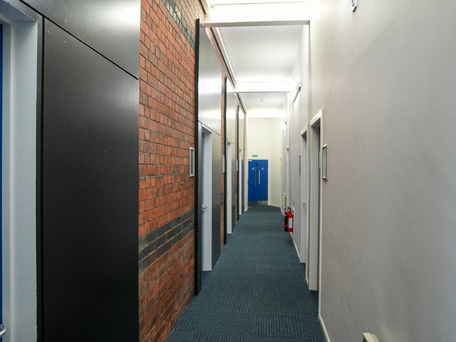 A narrow office corridor with an exposed brick wall on one side and office doors on the other
