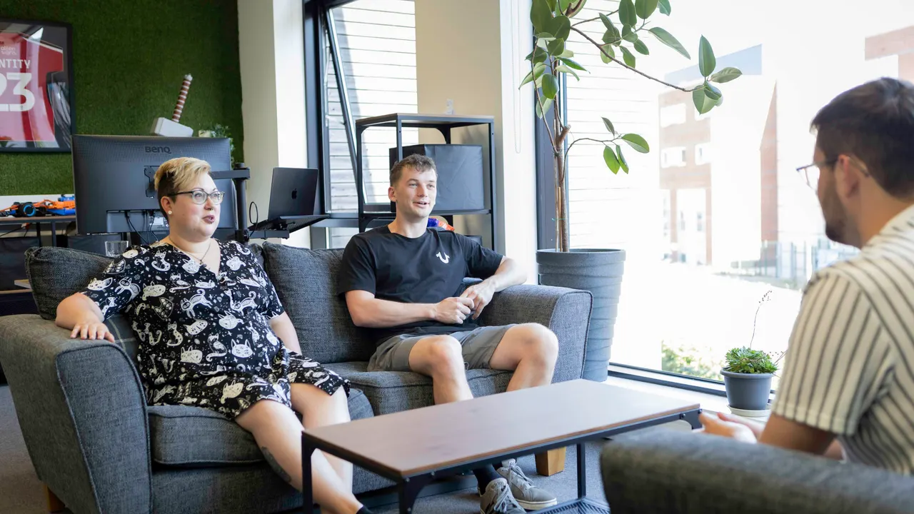 Three people talking on grey sofas in a bright office, beside a tall window and a potted plant