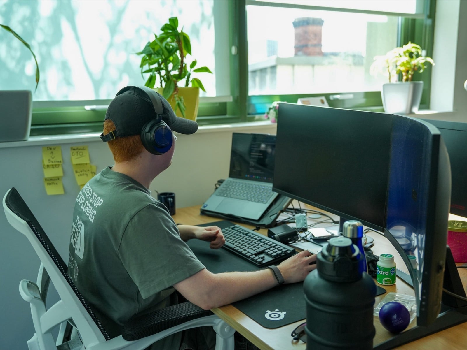 Someone in headphones working at a desk with a large monitor and laptop, sticky notes on the wall and plants on the windowsill