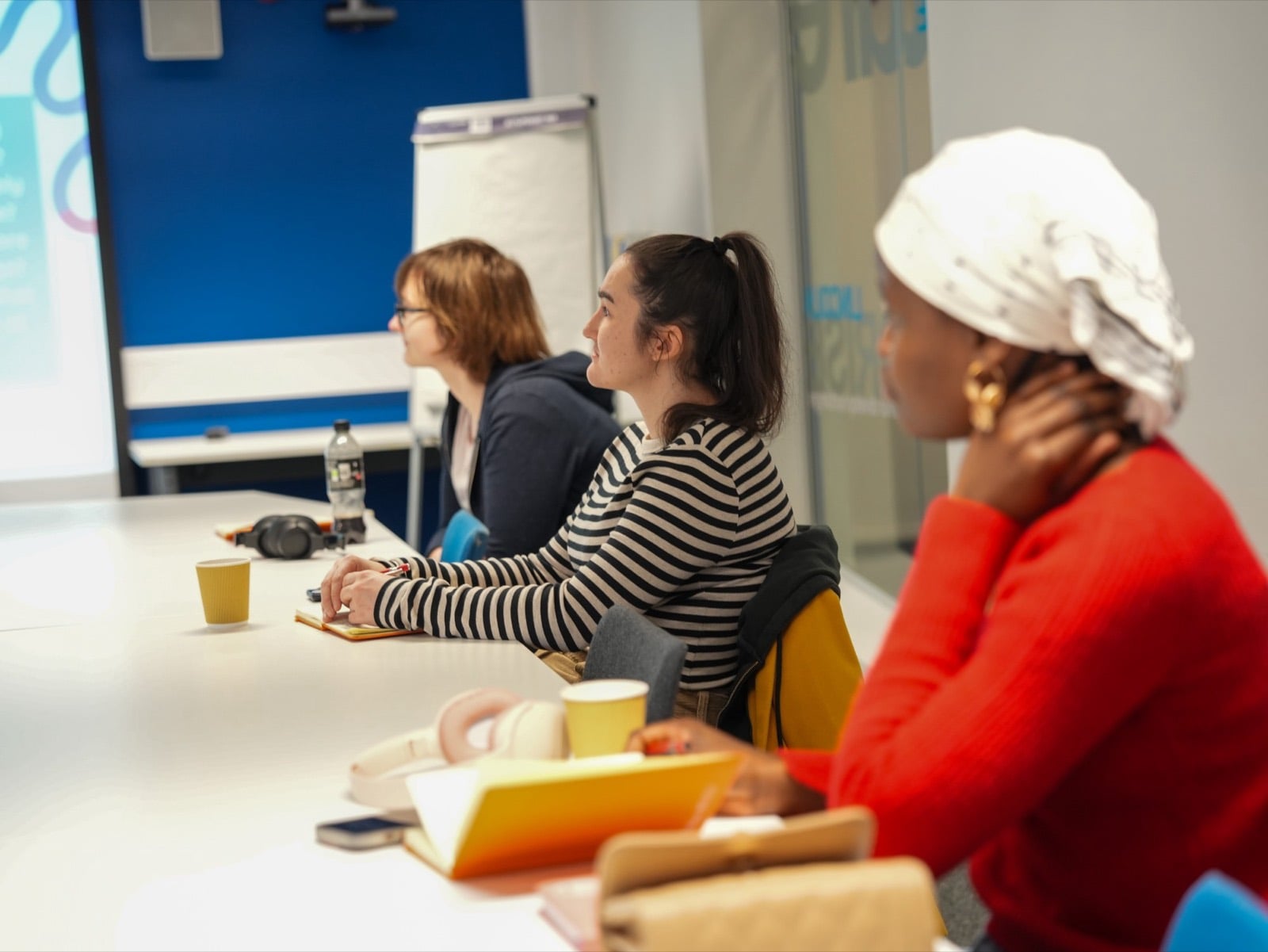 Three people at a workshop, listening with notebooks and coffee in front of a presentation screen