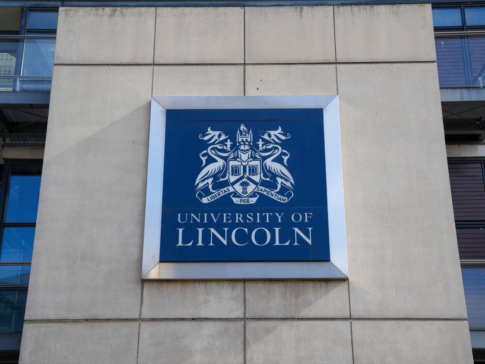 The University of Lincoln crest on a framed blue panel, mounted on a stone-clad campus building