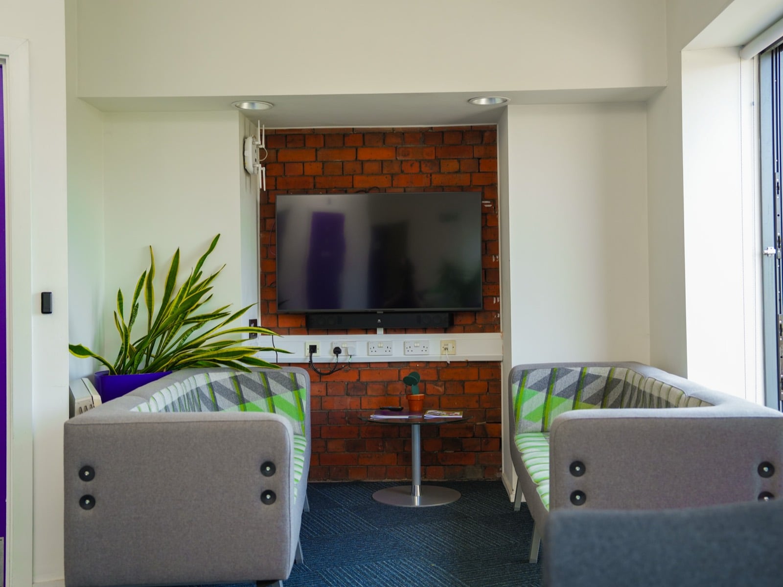 A breakout area with armchairs and a low table facing a wall-mounted screen on exposed brick