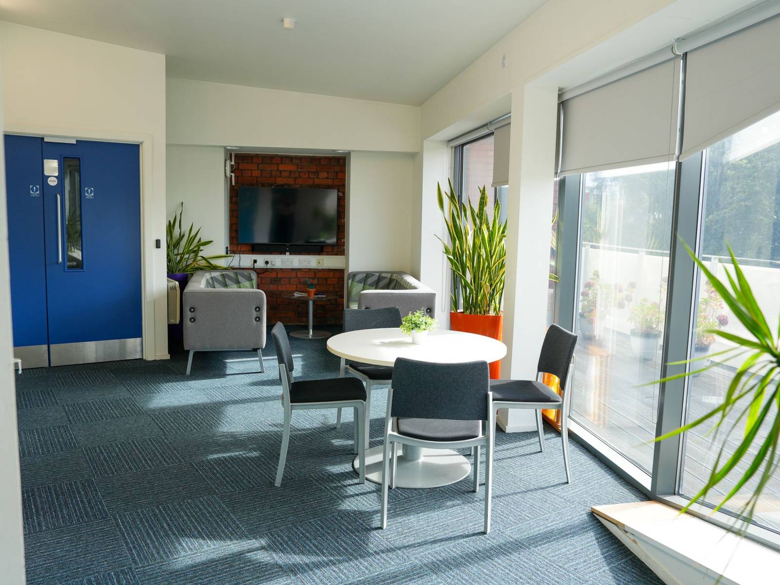 A breakout area with a round table, armchairs and a screen on exposed brick, beside glass doors onto a planted terrace