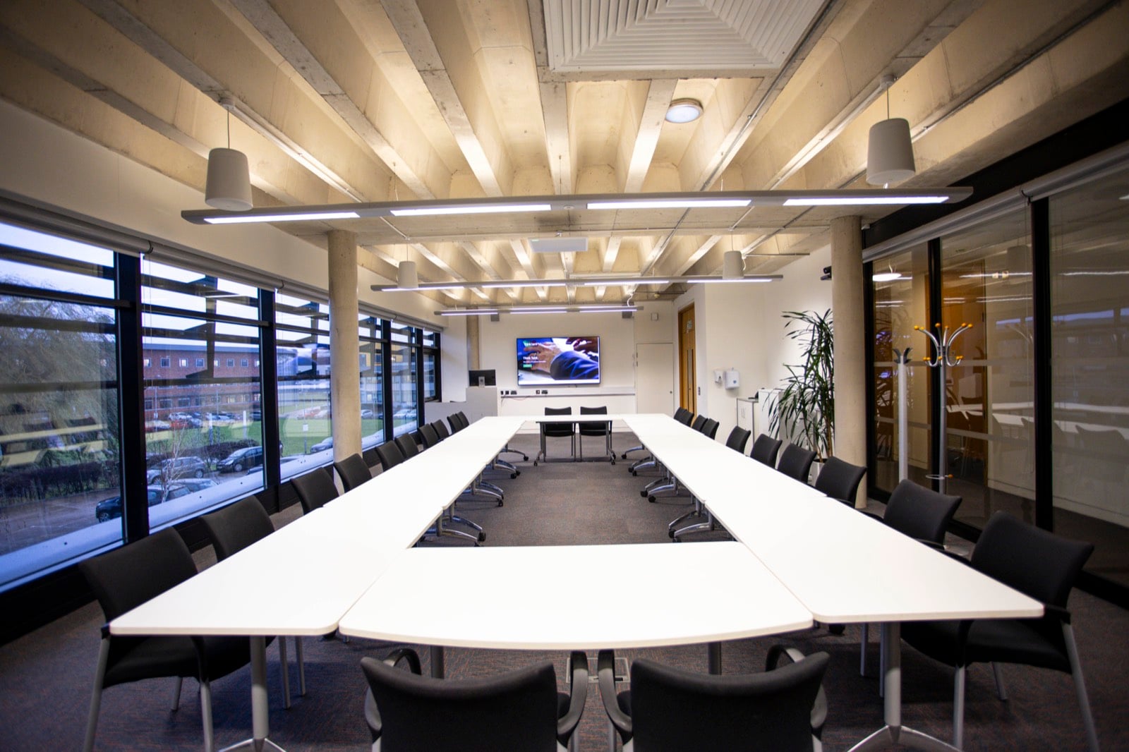 A long hollow-square boardroom under an exposed concrete ceiling, with a wall-mounted screen and windows onto the car park