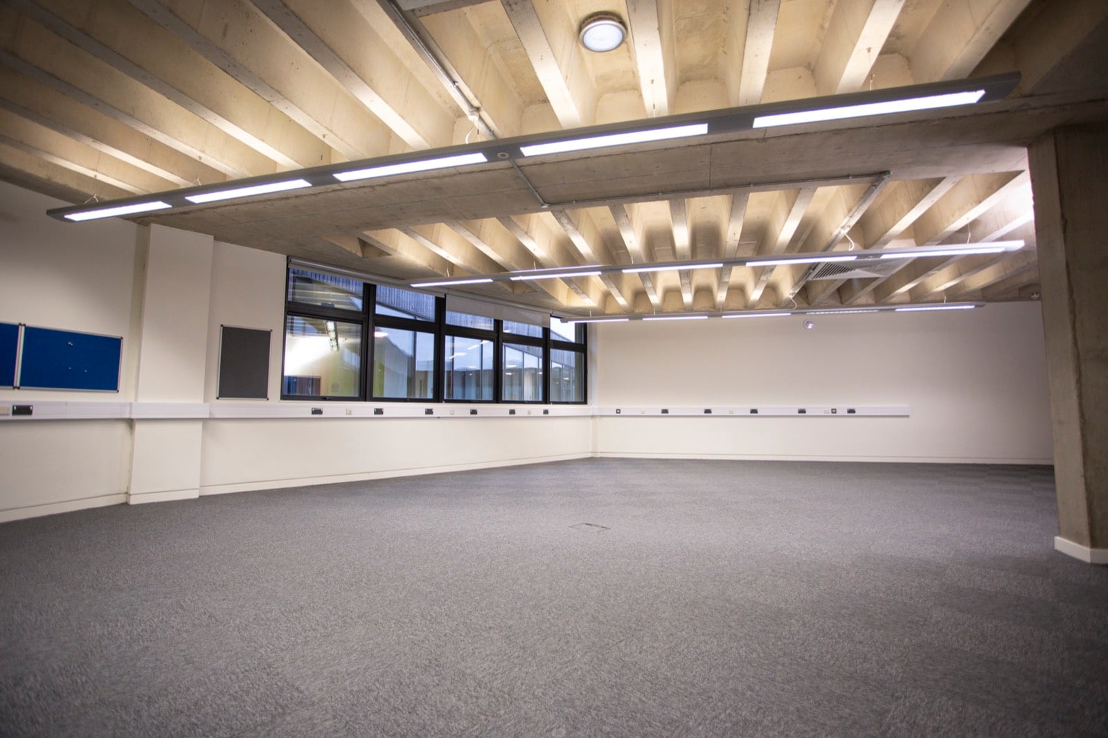 An empty carpeted office floor with a ribbon of windows and a run of power sockets along the wall
