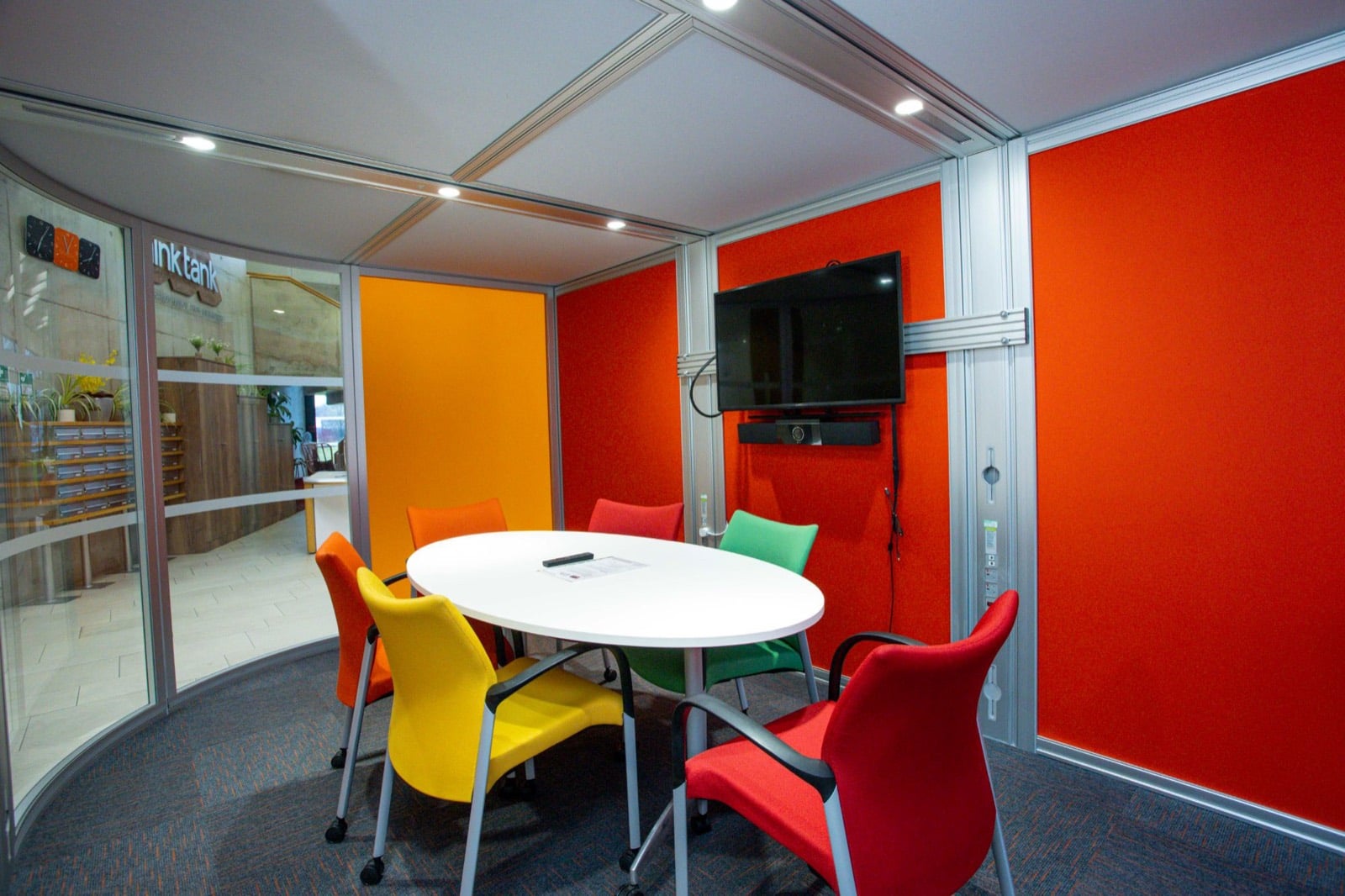 Inside a glass café pod at Think Tank, an oval table with coloured chairs, a wall-mounted screen and a view out to reception