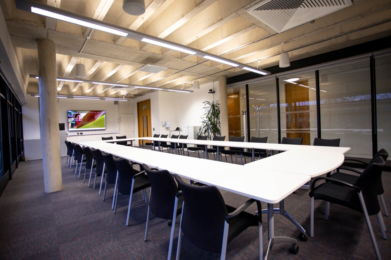 The Orange Room at Think Tank, a long hollow-square table under an exposed concrete ceiling, with extra theatre seating at the far end