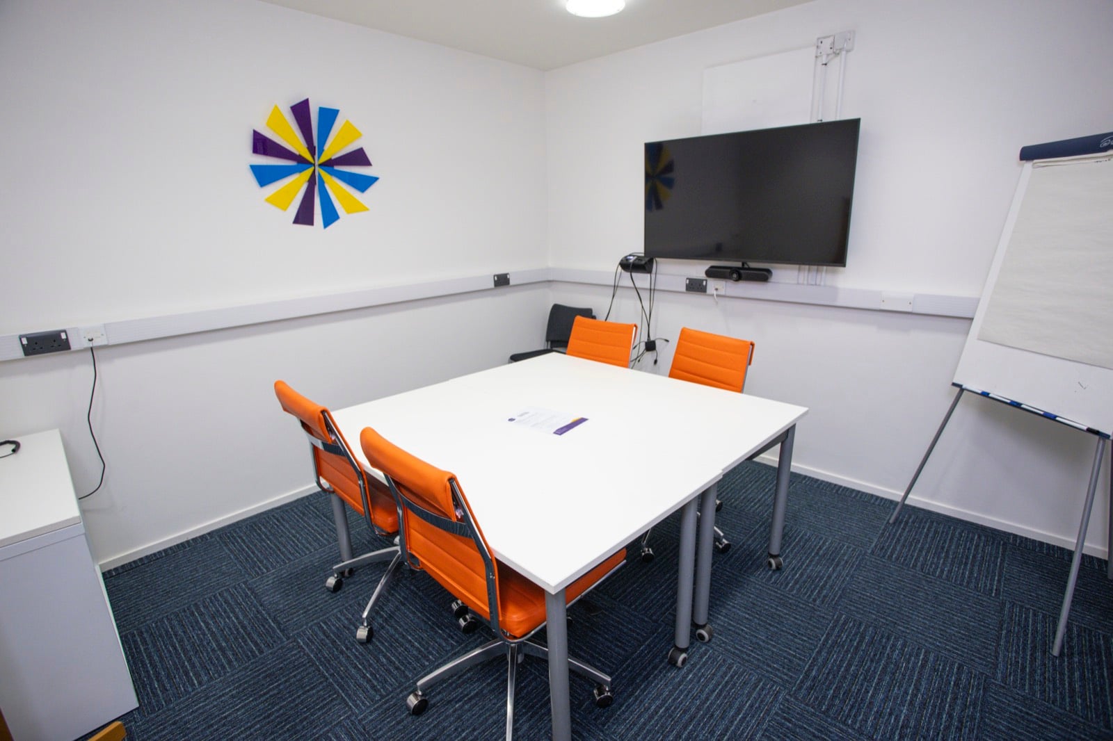 The Sparkhouse meeting room, with a square table, orange chairs, a wall-mounted screen and a flipchart