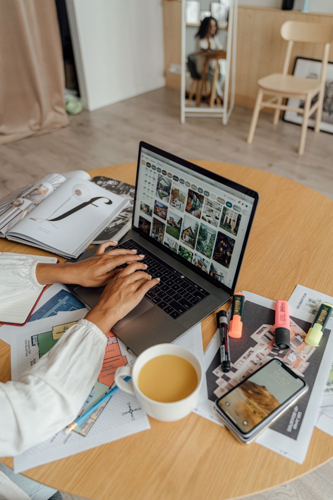 Someone working on a laptop at a round table covered in floor plans, highlighters, a phone and a mug of tea